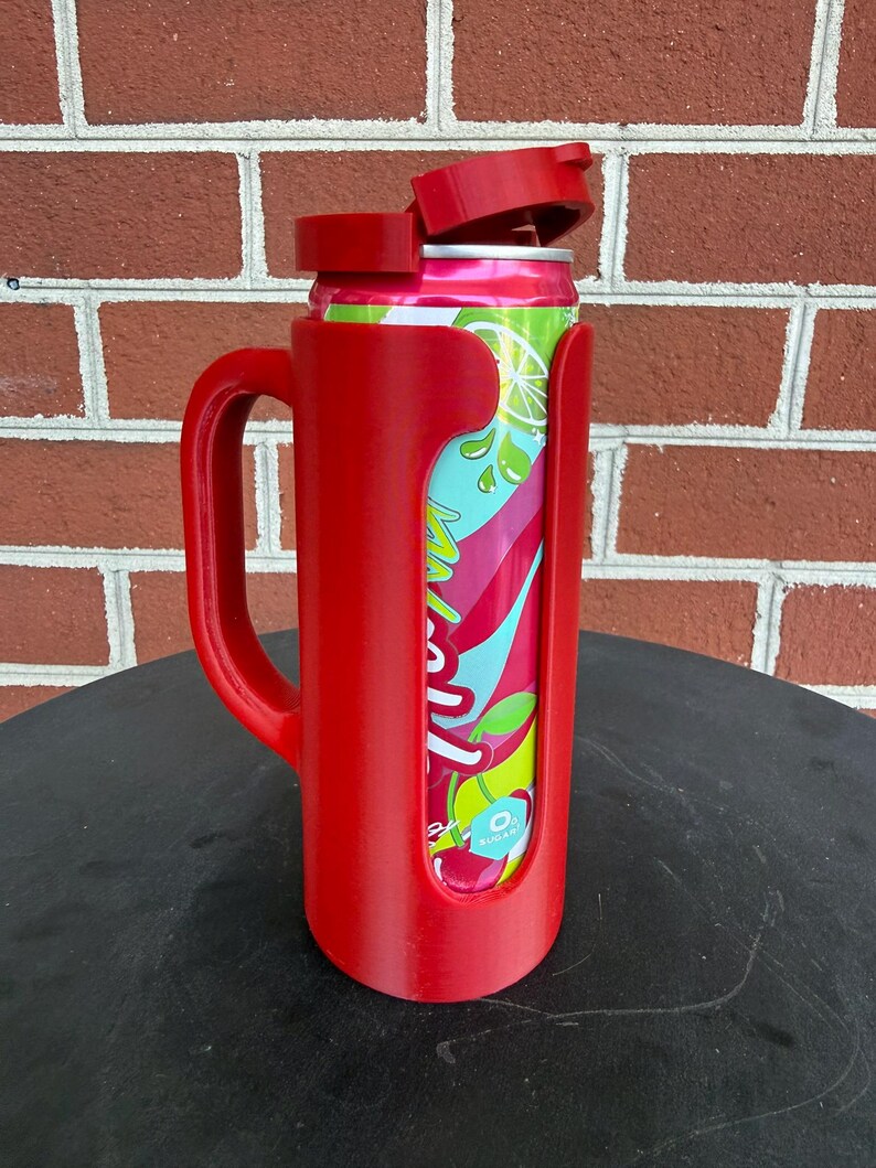 May include: A red plastic beverage can holder with a side handle and a hinged lid. The holder features a vertical cutout that reveals a colorful soda can with pink, teal, and lime green graphics, including a small white label reading 0g sugar.