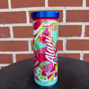 May include: A cylindrical Alani Nu energy drink can in Cherry Twist flavor, featuring a vibrant pink, red, and lime green graphic design with cherry and lime illustrations. The can is topped with a blue plastic lid and displays 12 fl oz text.