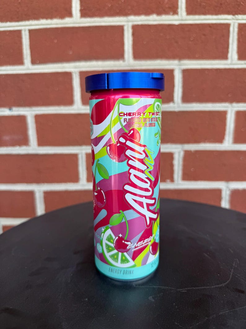 May include: A cylindrical Alani Nu energy drink can in Cherry Twist flavor, featuring a vibrant pink, red, and lime green graphic design with cherry and lime illustrations. The can is topped with a blue plastic lid and displays 12 fl oz text.