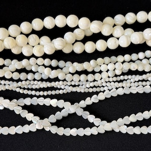 May include: Multiple strands of white mother-of-pearl beads arranged in rows. The top rows consist of round beads in varying sizes, while the bottom rows feature small heart-shaped beads strung together in a continuous, repeating pattern.
