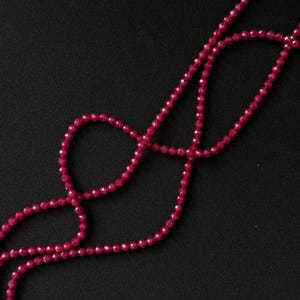 May include: A long strand of small, faceted, deep red beads arranged in a loose, looping pattern. Each individual bead features a multi-faceted surface that reflects light, creating a consistent, sparkling appearance along the entire length of the string.