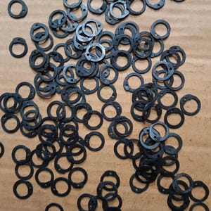Op de afbeelding: Een verzameling van talloze kleine, zwarte, cirkelvormige ringen, elk met een minuscuul gaatje nabij de rand. Deze onderdelen met een matte afwerking zijn verspreid over het kader en vertonen een uniforme vorm en consistente donkere kleur.