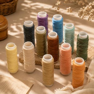 Könnte beinhalten: Eine Sammlung von zwölf zylindrischen Garnrollen in verschiedenen Farben, darunter Gelb, Creme, Beige, Orange, Pfirsich, Braun, Olivgrün, Dunkelgrün, Hellblau, Türkis, Dunkelgrau und Violett, jeweils mit einem weißen Kunststoffkern.