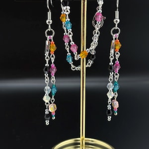 May include: A jewelry set consisting of a necklace and two matching dangle earrings. The pieces feature silver-tone chains adorned with multi-colored faceted crystal beads in shades of pink, blue, yellow, black, and clear, arranged in a pattern.