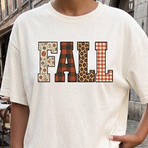 Peut inclure: Un t-shirt à manches courtes blanc cassé avec le mot FALL imprimé en grandes lettres majuscules sur la poitrine. Chaque lettre présente un motif distinct : le F est floral, le A est à carreaux rouges et verts, le L est léopard et le L est à carreaux orange.