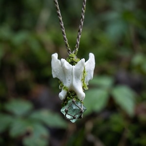 May include: A pendant featuring a white animal vertebra bone adorned with patches of green moss, suspended from a braided brown cord. A small, translucent, light green faceted crystal is secured at the base of the bone by a thin brown cord netting.