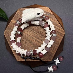 Può includere: Un bracciale con una testa di serpente bianca intagliata, perline bianche a forma di osso, perline rotonde rosa traslucide e piccoli distanziatori argentati a forma di teschio. Il design presenta una chiusura a cordino nero con pendenti.