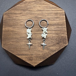 May include: A pair of silver-toned hoop earrings featuring dangling white bone vertebrae segments and a four-pointed star charm. The earrings are constructed with a circular hoop closure and a metallic finish, showcasing a combination of organic bone elements and polished metal components.