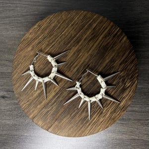 Op de afbeelding: Een paar oorringen bestaande uit een reeks gebroken witte, gesegmenteerde botachtige kralen afgewisseld met scherpe, zilverkleurige metalen punten. De ronde oorbellen zijn voorzien van een scharnierend sluitingsmechanisme aan de bovenzijde.