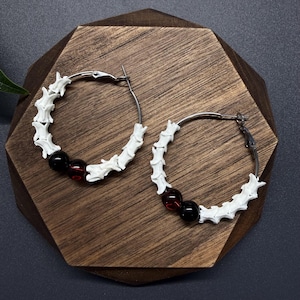 May include: A pair of hoop earrings featuring silver-toned metal wires adorned with white, segmented bone-like pieces and two smooth, round beads in deep red and black, arranged in a circular pattern along the lower half of each hoop.