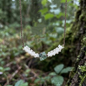 May include: A necklace featuring a brown cord strung with a series of small white vertebrae bones on each side, centered with a single translucent, multifaceted green and blue fluorite crystal held in a wire wrap setting.