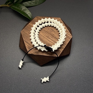 Könnte beinhalten: Ein Armband bestehend aus einer zweireihigen Kette aus cremefarbenen, strukturierten, knochenartigen Wirbelsegmenten. Das Schmuckstück wird mit einer schwarzen Kordel, einem verstellbaren Schiebeknoten und zwei passenden Perlen verschlossen.