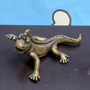 May include: A small, metallic brass-colored lizard figurine with a textured back, a long curved tail, and four legs. The lizard has a wide, open mouth and prominent eyes, with a polished finish that reflects light across its detailed, scaly surface.