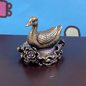 May include: A small, metallic, brass-colored figurine of a duck resting on a circular base decorated with sculpted floral elements. The duck has detailed wing feathers and a smooth head, while the base features textured, layered petals.