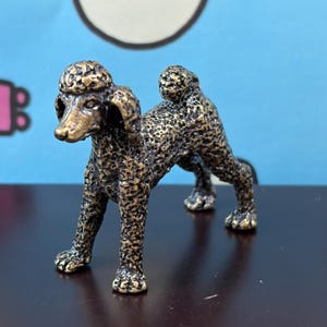 May include: A small, standing poodle figurine crafted from a dark, metallic material with a textured, bronze-toned finish. The sculpture features the dog's characteristic curly coat, a pom-pom tail, and detailed facial features, captured in a static pose.