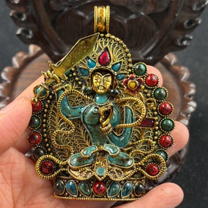May include: A circular gold-toned pendant featuring a central turquoise-coloured deity figure holding a sword. The piece is adorned with inlaid red and green stones, intricate filigree metalwork, and a decorative bail at the top for suspension.