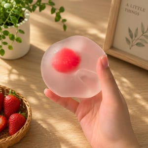 May include: A round, translucent, jelly-like dessert with a vibrant red centre. The object has a smooth, soft texture and a pale, semi-transparent appearance, resembling a mochi or fruit-filled confection, held up against a light-coloured background.