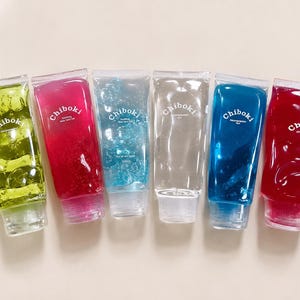 Könnte beinhalten: Sechs durchsichtige Kunststofftuben gefüllt mit farbigem, transluzentem Gel. Die Tuben sind in einer Reihe angeordnet und zeigen die Farben Limettengrün, Magenta, Hellblau, Transparent, Dunkelblau und Rot. Jede Tube ist mit dem weißen Text Chiboki beschriftet.