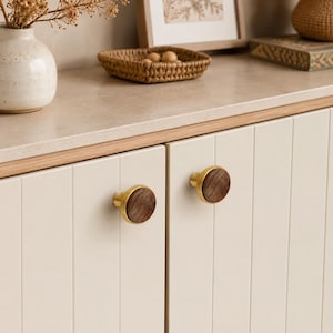 Può includere: Due pomelli circolari per armadietti con un inserto centrale in legno marrone scuro circondato da un bordo in metallo dorato spazzolato liscio. Questi componenti di ferramenta sono montati su ante di armadietti color bianco sporco.