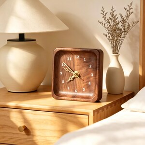 Könnte beinhalten: Eine quadratische Holzuhr mit dunkelbrauner Holzmaserung, weißen Ziffern von 1 bis 12 und goldfarbenen Metallzeigern. Die Uhr hat einen glatten, abgerundeten Rahmen und steht auf einer hellen Holzoberfläche.