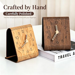 Peut inclure: Deux horloges de bureau en bois arquées avec des aiguilles dorées et des chiffres noirs. L'une est en bois clair avec un grain visible, l'autre en bois brun foncé. Le texte "Crafted by Hand", "Carefully Polished" et "Feel the temperature of time with your fingertips" est présent.