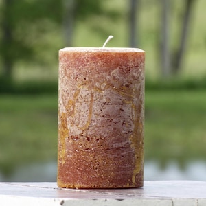 May include: A cylindrical pillar candle with a textured, mottled surface in shades of burnt orange, copper, and brown. The wax features a rustic, uneven finish with subtle metallic gold streaks running vertically along the sides and a single white wick.