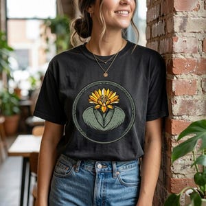 Könnte beinhalten: Ein schwarzes Kurzarm-T-Shirt mit einer kreisförmigen Grafik auf der Brust. Das Design zeigt eine stilisierte gelbe Blüte, die aus einer grünen Sukkulente wächst, eingefasst in einen dunkelgrünen Kreis mit geometrischen Linien.