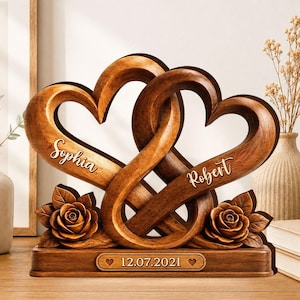 May include: A carved wooden sculpture featuring two interlocking hearts. The left heart is engraved with the name Sophia, and the right heart is engraved with the name Robert. Two carved roses sit at the base, which displays the date 12.07.2021.