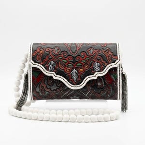 Puede incluir: Un bolso de mano rectangular de cuero negro con un intrincado patrón floral en relieve y detalles en rojo, verde y plata. Incluye una solapa festoneada, ribetes de cuero blanco, una correa de cuentas blancas y borlas de cadena metálica.