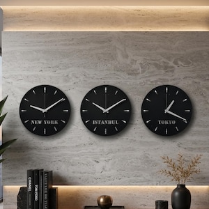 May include: Three circular black wall clocks with silver hands and markers. Each clock features a city name printed in silver text at the bottom: NEW YORK, ISTANBUL, and TOKYO. The clocks have a clean, simple design with no numbers.