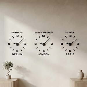 May include: Three black wall clocks arranged horizontally, each featuring numbers 3, 6, 9, and 12 with simple tick marks. The clocks are labeled Germany Berlin, United Kingdom London, and France Paris in black text above and below each clock.