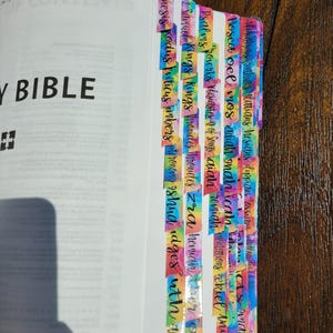 Könnte beinhalten: Eine Heilige Bibel mit bunten, im Tie-Dye-Muster gestalteten Kleberegistern an den Seitenrändern. Jedes Register trägt schwarzen kursiven Text, der verschiedene Bücher der Bibel benennt, darunter Genesis, Exodus, Levitikus, Numeri, Deuteronomium, Josua, Richter, Rut, Samuel, Könige, Chroniken, Esra, Nehemia, Ester, Hiob, Psalmen, Sprüche, Prediger, Hohelied, Jesaja, Jeremia, Klagelieder, Hesekiel, Daniel, Hosea, Joel, Amos, Obadja, Jona, Micha, Nahum, Habakuk, Zefanja, Haggai, Sacharja, Maleachi, Matthäus, Markus, Lukas, Johannes, Apostelgeschichte, Römer, Korinther, Galater, Epheser, Philipper, Kolosser, Thessalonicher, Timotheus, Titus, Philemon, Hebräer, Jakobus, Petrus, Johannes, Judas und Offenbarung.