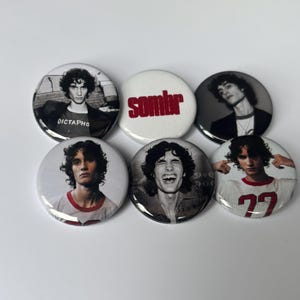 Può includere: Un set di sei spille rotonde. Cinque spille presentano ritratti fotografici in bianco e nero di una persona con capelli ricci, una delle quali include il testo "DICTAPHO" e un'altra "77". Una spilla bianca presenta il testo rosso "sombr".