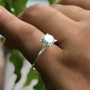 May include: A silver ring featuring a central round white stone surrounded by a halo of small green stones. The ring has a thin, polished silver band and is worn on a finger, showcasing the circular setting and the contrast between the stones.