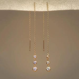 Puede incluir: Un par de pendientes de hilo de oro, cada uno con una cadena larga y delicada que suspende tres piedras redondas, transparentes y facetadas en disposición vertical. Los pendientes tienen un poste recto y fino en el extremo superior.
