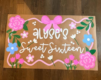 Könnte beinhalten: Ein rechteckiges Schild aus braunem Papier mit dem weißen handgeschriebenen Text Alyse's Sweet Sixteen. Das Design ist von einem wellenförmigen rosa Rand mit einer großen rosa Schleife oben umrahmt und mit gemalten rosa und blauen Blumen sowie grünen Blättern verziert.