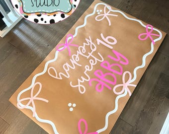Könnte beinhalten: Ein rechteckiges, hellbraunes Schild mit weißem Wellenrand und rosa Schleifen an den Ecken. Der Text "happy sweet 16 ABBY" ist in hellrosa und leuchtend rosa Schreibschrift gehalten. Das Design enthält kleine weiße Punkte als Akzente.