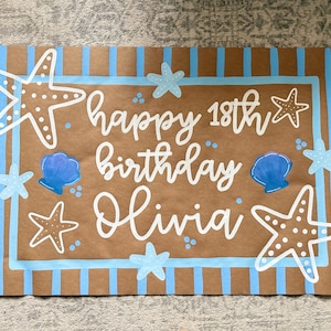 Könnte beinhalten: Braunes Papierschild mit blauen Streifen und blauem Rand. Auf dem Schild steht "Happy 18. Geburtstag Olivia" in weißer Schrift. Verziert mit weißen Seesternen und blauen Muscheln.