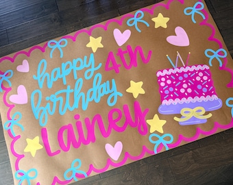 Könnte beinhalten: Ein braunes Papier-Geburtstagsschild mit den Worten "happy 4th birthday Lainey" in Blau und Pink. Das Schild ist mit rosa Herzen, gelben Sternen und blauen Schleifen verziert. Ein rosa Kuchen mit Kerzen ist ebenfalls abgebildet.