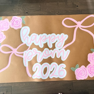 Puede incluir: Un cartel marrón con las palabras "happy prom 2026" en letras blancas y rosas. El cartel está decorado con lazos y rosas rosas. Las rosas tienen hojas verdes. El cartel está sobre un suelo de madera.