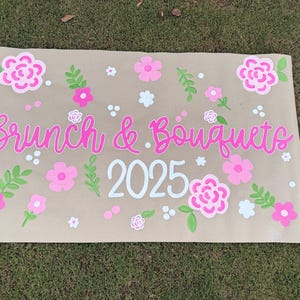 May include: A tan sign with the words "Brunch & Bouquets 2025" in pink script. The sign is decorated with pink, white, and green floral designs, including flowers and leaves. The sign is on a grassy surface.