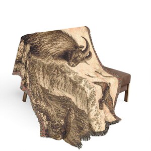 May include: A woven throw blanket featuring a detailed illustration of a mountain goat standing on a rocky cliffside with a bird flying in the sky. The design uses shades of brown, tan, and cream with fringed edges along the sides and bottom.
