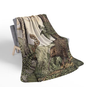 May include: A throw blanket featuring a detailed illustration of a brown kiwi bird standing in a lush forest environment with green ferns, textured tree trunks, and a pale sky background, draped over a grey chair with light wooden legs.