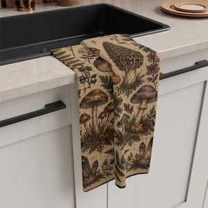 May include: A rectangular kitchen towel featuring a detailed botanical print on a beige background. The pattern displays various types of mushrooms, including morels, alongside fern leaves, small wildflowers, and pointed crystal formations.
