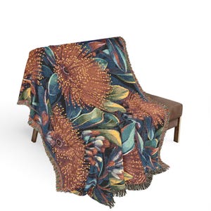 May include: A woven throw blanket featuring a dark blue background with a large-scale botanical pattern of rust-coloured protea flowers and green leaves. The blanket has a textured, tapestry-like weave and finished edges with short, frayed fringe detailing.