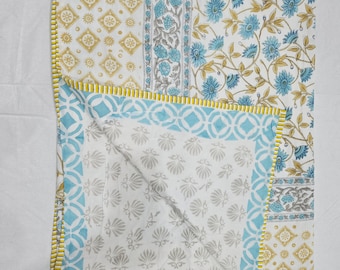 May include: A folded cotton quilt featuring a white base with a multi-patterned design. The top section displays yellow geometric shapes and blue floral motifs. The underside shows a light blue circular pattern and gray shell-like prints.
