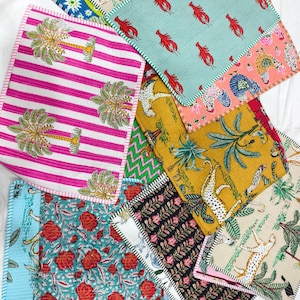 May include: A collection of rectangular quilted fabric placemats featuring various patterns including pink and white stripes with palm trees, red lobsters on light blue, yellow with leopards, and floral prints in teal, black, and cream colours.