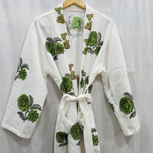 Puede incluir: Una bata blanca con textura de gofre que presenta un estampado floral repetitivo en tonos verde y gris oscuro. La prenda incluye mangas largas, cuello de chal y un cinturón de tela a juego en la cintura, todo exhibido en una percha de madera.
