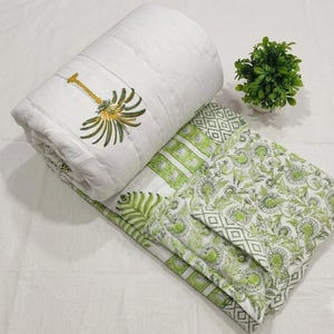 May include: A white quilted blanket featuring a green and yellow embroidered palm tree motif on the rolled section, with a green floral and geometric block print pattern on the unfolded portion, paired with a small potted green plant.