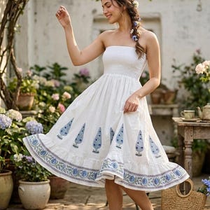 May include: A white strapless midi dress featuring a smocked bodice and a gathered skirt. The hem is decorated with a blue floral border print, while the main skirt displays a repeating pattern of blue paisley-style motifs on the white fabric.